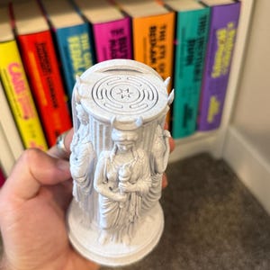 May include: A white, speckled, cylindrical sculpture featuring three figures in draped robes standing around a central column. Each figure wears a crescent moon headpiece and holds a torch. The top of the column displays a circular labyrinth pattern.