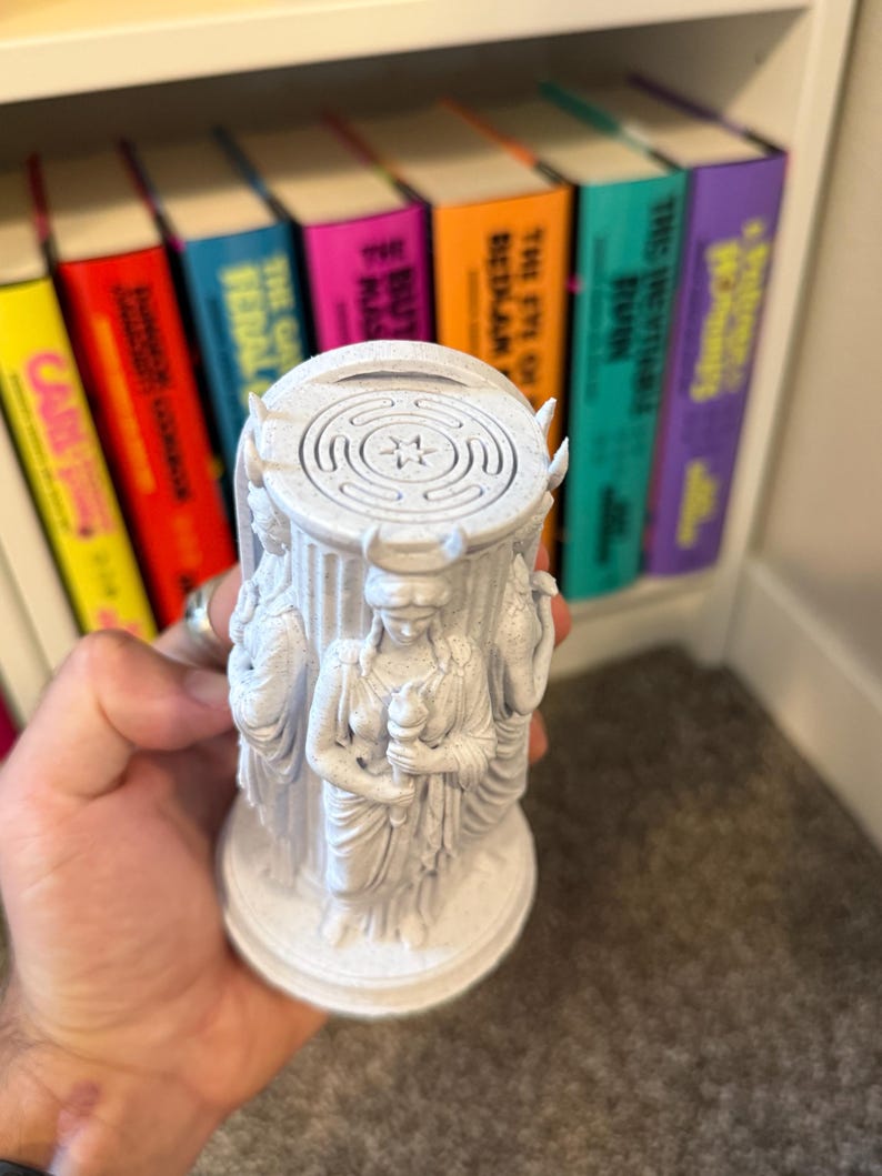May include: A white, speckled, cylindrical sculpture featuring three figures in draped robes standing around a central column. Each figure wears a crescent moon headpiece and holds a torch. The top of the column displays a circular labyrinth pattern.