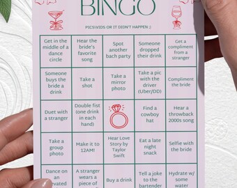 May include: A pink and white Bachelorette Bingo card featuring fun activities. The card includes prompts such as "Take a mirror photo" and "Hear a throwback 2000s song". The card is held by a person.