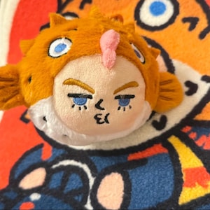 Puede incluir: Un peluche con forma de pez globo con cuerpo naranja y rostro con ojos azules y un acento rosa. El juguete tiene un cuello blanco y está sobre un fondo estampado colorido.