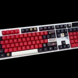 Könnte beinhalten: Eine weiße mechanische Tastatur mit roten und schwarzen Tastenkappen. Die Tastatur verfügt über ein individuelles Design mit dem Text "44 RADIO" und "FERRARI" sowie einer Rennwagen-Grafik. Die Tastatur hat eine weiße Leertaste und einen Lautstärkeregler.