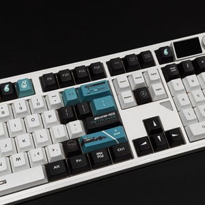 May include: A white mechanical keyboard with black and teal keycaps. The keyboard has a number pad, function keys, and a small display screen. The keycaps feature symbols and text, including "AMG PETRONAS".