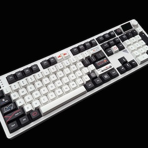 Puede incluir: Un teclado mecánico blanco y negro con teclas personalizadas. El teclado presenta una mezcla de teclas blancas y negras, con algunas teclas que muestran logotipos y texto. El teclado está sobre un fondo negro.