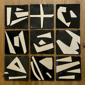 May include: Nine square black coasters with abstract white geometric designs. The coasters are arranged in a 3x3 grid, showcasing a variety of shapes and patterns. The coasters are displayed on a wooden surface.