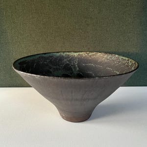 Puede incluir: Un cuenco de cerámica marrón oscuro hecho a mano con forma acampanada. El borde interior presenta un esmalte moteado verde y negro, que contrasta con el exterior. El cuenco está sobre una superficie blanca contra un fondo verde oscuro.