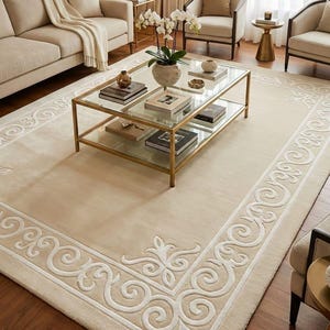 May include: A cream-coloured area rug with a white, ornate border and a gold-framed glass coffee table. The rug features a detailed scroll and floral design. The room has a neutral colour palette with beige furniture.