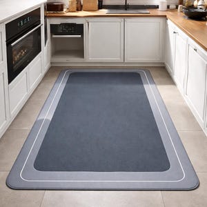 May include: A rectangular kitchen rug in shades of gray and blue. The rug has a darker gray center, a lighter gray border, and a thin white outline. The rug is placed in front of white kitchen cabinets and a black oven.