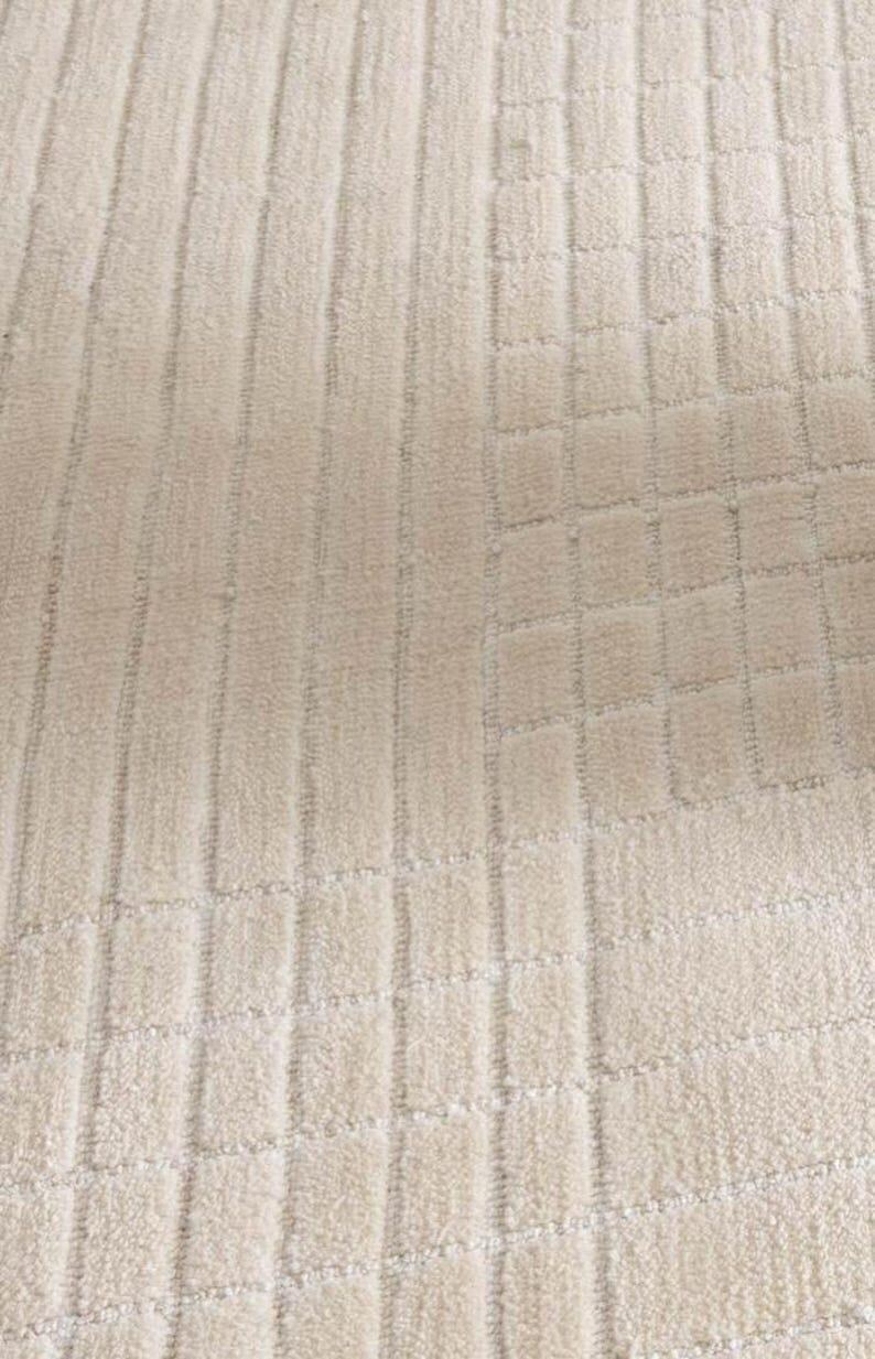 Pu&ograve; includere: Primo piano di un tessuto beige strutturato con un motivo ripetuto di linee verticali e blocchi quadrati. Il tessuto ha un aspetto morbido e felpato, che suggerisce un tappeto o un tessuto.