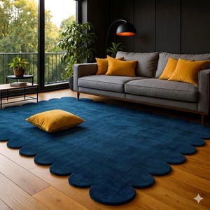May include: A modern living room featuring a large, scalloped-edge, deep blue rug. A gray sofa with mustard-colored pillows sits against a dark wall. A matching mustard-colored square pillow rests on the rug. The room has hardwood floors and a large window.
