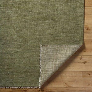 May include: A green rug with a textured surface. The corner of the rug is folded back, revealing the underside with a lighter color and a fringed edge. The rug is placed on a wooden floor.