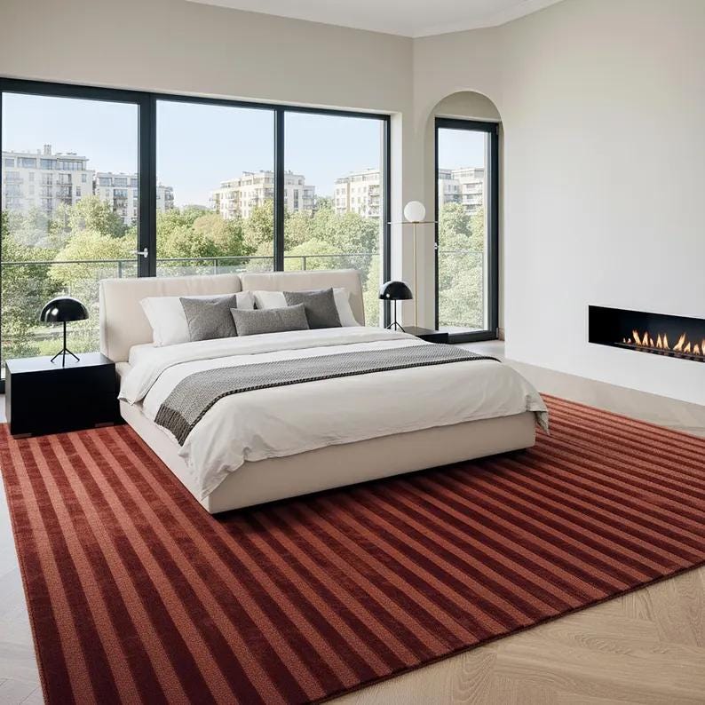 Pu&ograve; includere: Una camera da letto moderna con un letto grande, un tappeto rosso a righe e ampie finestre con vista su un paesaggio urbano. Il letto ha lenzuola bianche e cuscini grigi. Due lampade nere sono sui comodini. Un camino &egrave; sul muro.