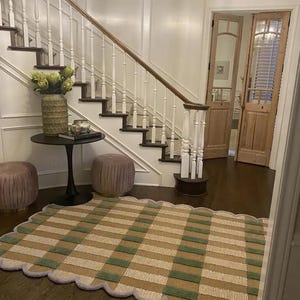May include: A patterned rug with a scalloped edge, displaying stripes of tan, green, and off-white. The rug is laid on a dark wooden floor in a bright hallway, with a staircase and decorative items visible in the background.