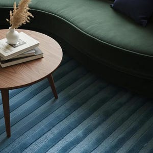 Pu&ograve; includere: Un tavolino rotondo in legno con tre gambe, con libri e un piccolo vaso bianco con piante essiccate. Un divano curvo verde scuro e un tappeto blu a righe sono sullo sfondo.