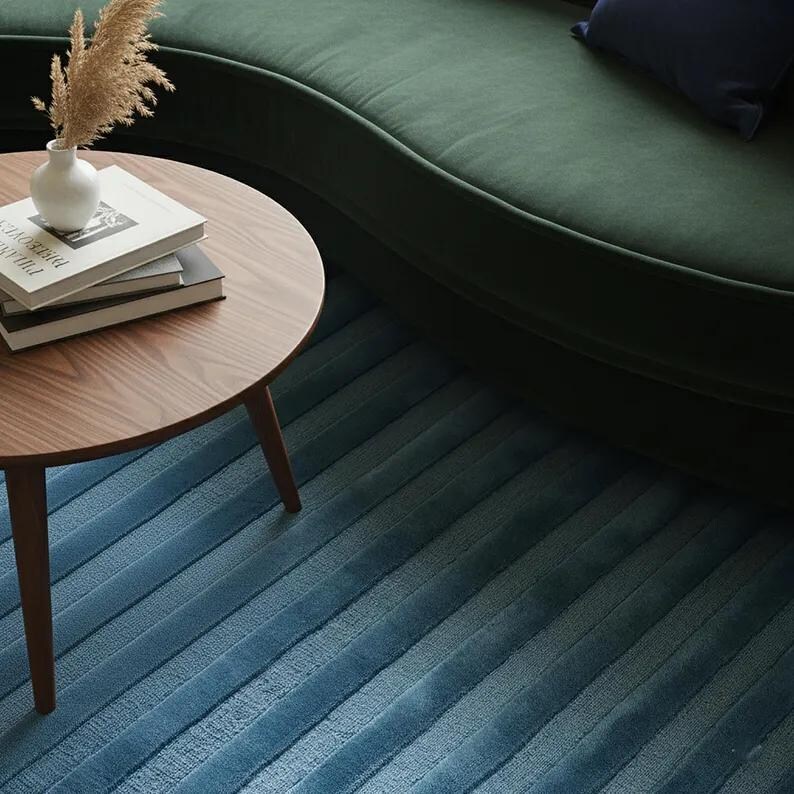 Pu&ograve; includere: Un tavolino rotondo in legno con tre gambe, con libri e un piccolo vaso bianco con piante essiccate. Un divano curvo verde scuro e un tappeto blu a righe sono sullo sfondo.