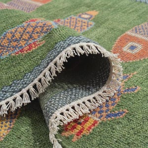 May include: Close-up of a rolled-up green rug with a geometric pattern. The rug features a mix of colors, including orange, blue, and red, with a cream-colored fringe along the edge. The rug's design is visible on the unrolled portion.