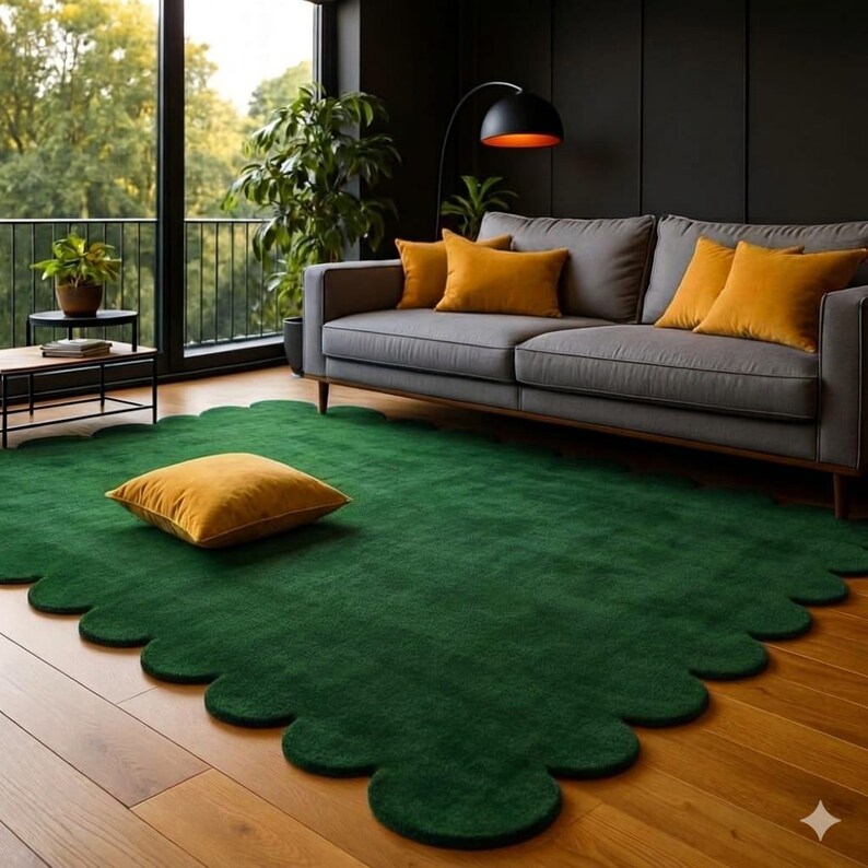 May include: A green, scalloped-edge rug with a plush texture dominates the foreground. A gray sofa with mustard-colored pillows sits against a dark wall. A mustard-colored square pillow rests on the rug. The room has hardwood floors and a large window.