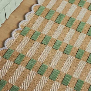 May include: A patterned rug featuring a design of tan, white, and green rectangles. The rug has a scalloped edge with a light grey border. It is placed on a wooden floor, with a section of a green sofa visible in the background.
