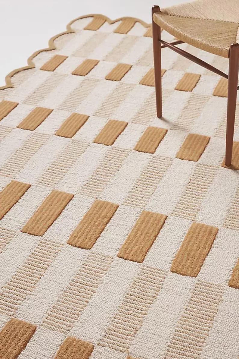 May include: A beige and white patterned rug with a scalloped edge. The rug features alternating rows of beige rectangular blocks and white striped sections. A wooden chair leg is visible in the upper right corner.