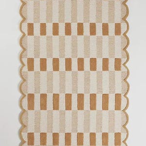 May include: A rectangular rug with a scalloped edge. The rug features a repeating pattern of vertical stripes in alternating shades of beige and cream. The rug's border is a solid beige colour.