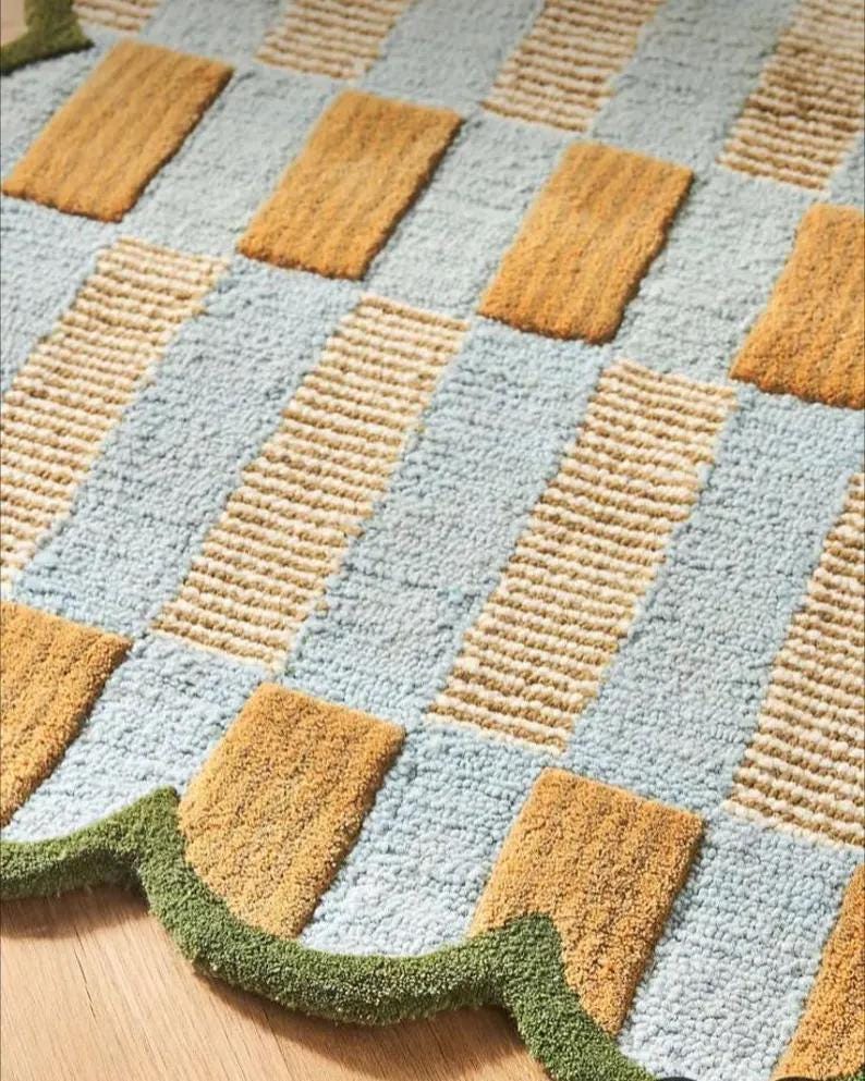 May include: A close-up of a textured rug with a geometric pattern. The rug features alternating light blue and tan rectangular blocks, with a green scalloped edge. The rug's design is a mix of solid and striped textures.