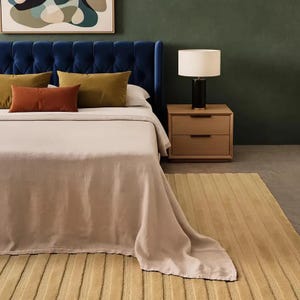 Pu&ograve; includere: Una scena di camera da letto con una testiera blu trapuntata, un letto con una coperta rosa chiaro e cuscini marroni e verdi. Un comodino in legno con una lampada e un tappeto giallo completano la scena.