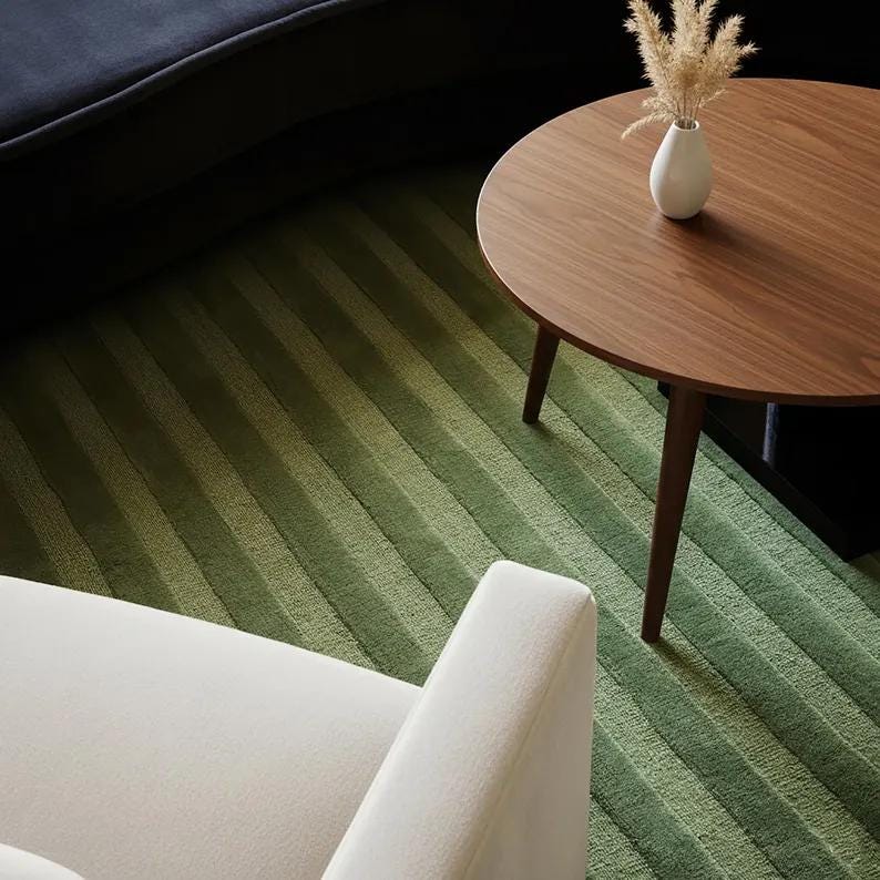 Pu&ograve; includere: Un moderno salotto con un tavolino rotondo in legno, un piccolo vaso bianco con piante secche, un tappeto verde a righe e una poltrona bianca. Le strisce del tappeto sono diagonali.
