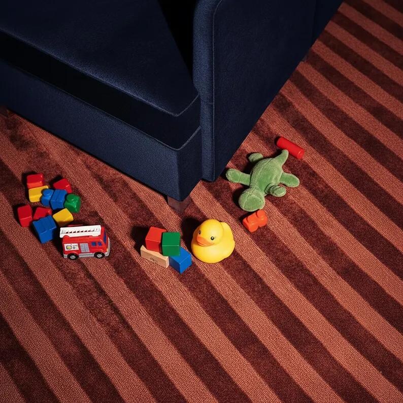 Pu&ograve; includere: Una collezione di giocattoli per bambini sparsi su un tappeto a righe. I giocattoli includono un camion dei pompieri rosso, una papera di gomma gialla, una rana di peluche verde e blocchi colorati. Un divano blu navy &egrave; sullo sfondo.