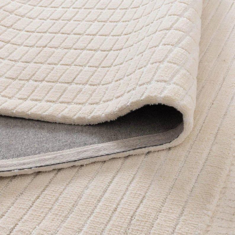 Pu&ograve; includere: Primo piano di un tappeto color crema. Lo strato superiore presenta un motivo geometrico a rombi, mentre quello inferiore ha un design a coste. Il tappeto &egrave; arrotolato, rivelando un retro grigio.