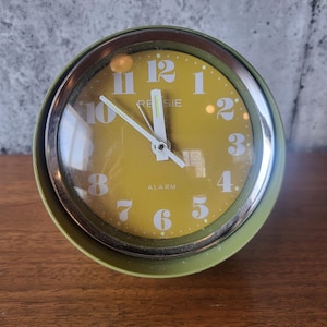 Peut inclure: Un réveil vert vintage avec un cadran jaune. L'horloge a des chiffres et des aiguilles blancs, avec le mot "RE SIE" imprimé au-dessus du centre. Le mot "ALARM" est imprimé en dessous. L'horloge est posée sur une surface en bois.