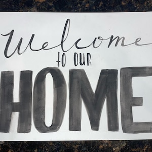 May include: A white rectangular sign with the words "Welcome to our Home" written in black ink. The word "Welcome" is in cursive, and "to our HOME" is in block letters.