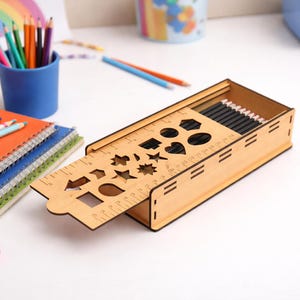 May include: A wooden pencil case with a ruler and shape cutouts, open to reveal pencils. Colorful pencils and notebooks are nearby. A pink marker and a drawing of a rainbow are also visible.