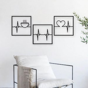 May include: Three black metal wall art pieces. One features a coffee cup with steam, another a heartbeat line, and the third a heart within a heartbeat line. Each is framed in a square. The art is displayed above a white chair.