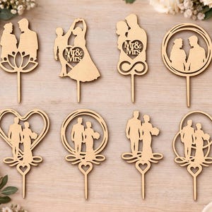 Puede incluir: Una colección de adornos de madera para tartas de boda en varios diseños. Siluetas de parejas se presentan en formas de corazón, círculo e infinito. Algunos adornos incluyen el texto "Mr & Mrs". Los adornos son de color marrón claro.