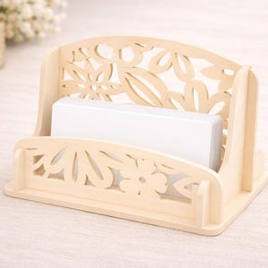 Può includere: Un portacarte in legno chiaro con un design floreale traforato. Il supporto ha una base rettangolare e un retro con una parte superiore curva. Una pila di carte bianche si trova all'interno.