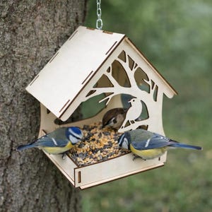 May include: A wooden bird feeder with a tree and bird silhouette design, hanging from a chain. Two blue and yellow birds are perched on the feeder, eating seeds. The feeder is attached to a tree trunk.