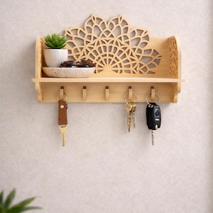 Puede incluir: Un llavero de madera de color claro con un diseño decorativo cortado con láser. Cuenta con un pequeño estante con una planta, un cuenco y ganchos para llaves. Las llaves y una llave de coche cuelgan de los ganchos. El estante está montado en la pared.