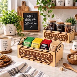 Può includere: Una cassetta portaoggetti in legno chiaro con la scritta COFFEE & TEA tagliata al laser sul davanti, riempita con varie bustine di tè e pacchetti di caffè. La cassetta presenta maniglie intagliate sui lati e un motivo floreale sotto il testo.