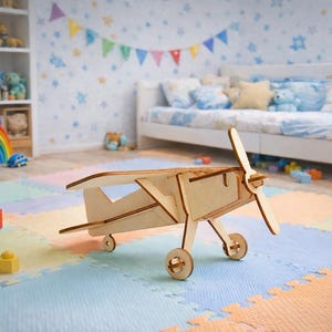 May include: A wooden toy airplane with a propeller and wheels sits on a colorful play mat. The plane is made of light-colored wood and has a classic design. The background includes a bed, toys, and a bookshelf, suggesting a child's room.