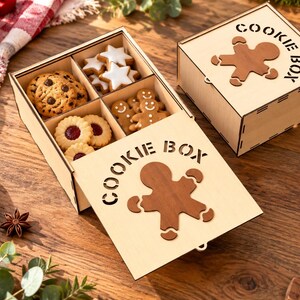 May include: A light-colored wooden box with four internal compartments filled with various cookies, including chocolate chip, star-shaped, and gingerbread figures. The lid features a cutout gingerbread man and the text COOKIE BOX.