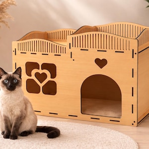 May include: A light-coloured wooden cat house with a heart-shaped window and a heart cutout. A Siamese cat with blue eyes sits on a white rug in front of the house. The cat house has a rounded roof design.