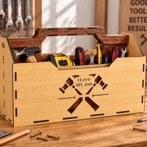 Peut inclure: Un porte-outils en bois clair avec une poignée en bois foncé, présentant une découpe laser de deux marteaux croisés et le texte "I Love MY Job" sur le panneau avant. La boîte ouverte contient divers outils à main, dont un marteau et un mètre ruban.