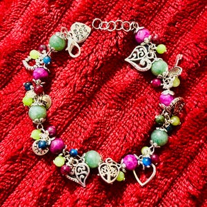 May include: A colourful beaded bracelet featuring green, purple, blue, and yellow beads. The bracelet includes silver charms, such as hearts and a tree of life design. The bracelet is displayed on a red textured background.