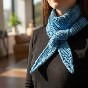 May include: A light blue knitted scarf is tied around the neck. The scarf is made of a textured knit fabric and is a solid light blue colour. The scarf is worn over a black long-sleeved top.