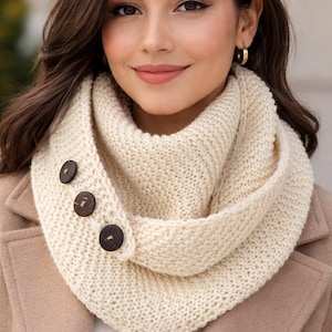 Könnte beinhalten: Cremefarbener Strickschal mit drei dunkelbraunen Knöpfen. Der Schal ist aus dickem, strukturiertem Garn gefertigt und wird um den Hals gelegt. Der Hintergrund zeigt eine Frau in einem beigen Mantel.