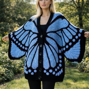 Könnte beinhalten: Eine blau-schwarze Häkelstrickjacke mit einem Schmetterlingsflügel-Design. Der Cardigan mit offenem Frontteil hat einen V-Ausschnitt, einen Knopfverschluss und lange Ärmel. Die Schmetterlingsflügel sind hellblau mit schwarzen Umrissen und Akzenten.