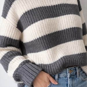 Könnte beinhalten: Ein grau-weiß gestreifter Pullover mit Rundhalsausschnitt. Der dick gestrickte Pullover hat breite horizontale Streifen und lange Ärmel. Die Person trägt eine blaue Jeans mit einer Hand in der Tasche.