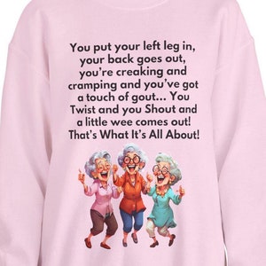May include: Pink sweatshirt with a humorous saying about getting older and a cartoon image of three older women laughing and dancing. The text reads: "You put your left leg in, your back goes out, you're creaking and cramping and you've got a touch of gout... You Twist and you Shout and a little wee comes out! That's What It's All About!"