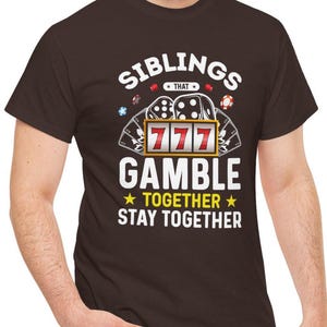 May include: A dark brown t-shirt with the text "SIBLINGS THAT GAMBLE TOGETHER STAY TOGETHER" in white. The design includes a slot machine with the number 777 and casino chips.