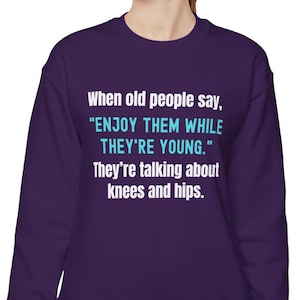 May include: Purple sweatshirt with a white and teal blue graphic print that says "When old people say, 'ENJOY THEM WHILE THEY'RE YOUNG.' They're talking about knees and hips."