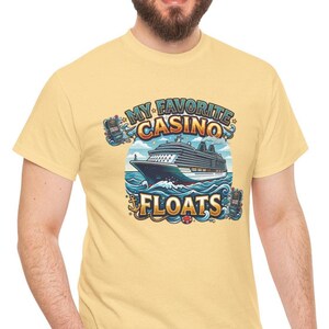 May include: A light yellow t-shirt featuring a colorful graphic of a cruise ship sailing on blue waves. The text "MY FAVORITE CASINO FLOATS" is displayed above and below the ship. The design includes casino-themed elements.